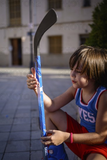 A young child sits on a paved street, holding a vibrant blue hockey stick. The scene captures a moment of quiet focus and passion for the sport in an urban environment