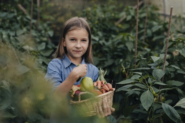A young girl in a blue shirt joyfully holds a basket filled with freshly harvested vegetables, surrounded by green foliage in a flourishing greenhouse setting