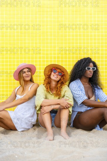 Three young women wearing summer clothes and sunglasses are sitting on the sand against a yellow tiled wall
