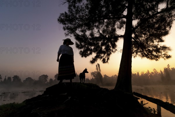 A serene silhouette of a woman and her Xoloitzcuintle dog at sunrise, against the backdrop of the tranquil waters and lush surroundings of Xochimilco, Mexico