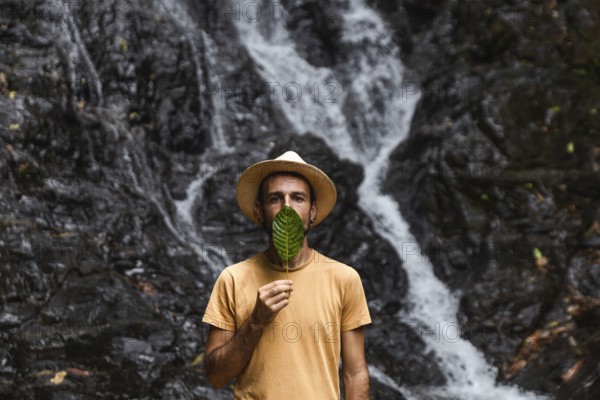Man from Spain holding a leaf in front of his face, with a waterfall in the Choco Andino rainforest, Ecuador. He is engaged in exploring the rich biodiversity and experiencing ecotourism