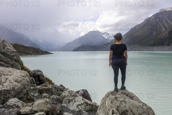 A woman stands on a rocky outcrop, taking in the serene autumn beauty of Mount Cook in New Zealand. The stunning lake and mountains form a breathtaking backdrop