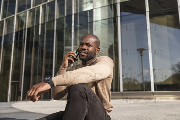 A cuban man sits outside a sleek, modern glass building, engaging in a conversation on their smartphone. The sunny ambiance adds warmth to the professional setting