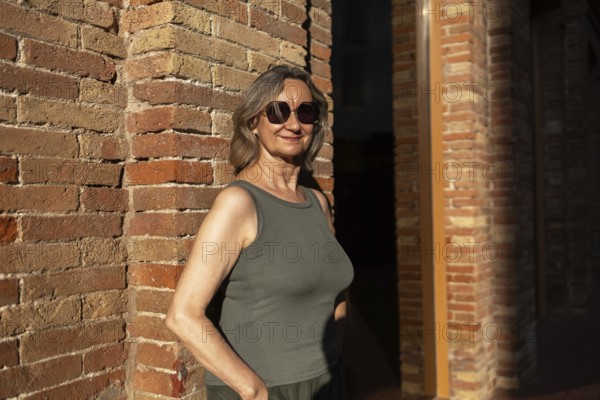 A joyful mature woman in casual attire stands against a brick wall, basking in the warm sunlight. Her smile and sunglasses reflect a relaxed, confident lifestyle