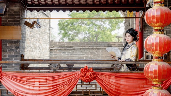 A woman in traditional Qing Dynasty attire, holding a fan, stands gracefully on a decorated balcony in Pingyao, China. The vibrant red lanterns frame the historical scene