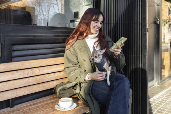 A woman enjoys a coffee while sitting on a bench outside a cafe, holding her phone and smiling at her small chihuahua on her lap. Sunlight creates a warm ambiance