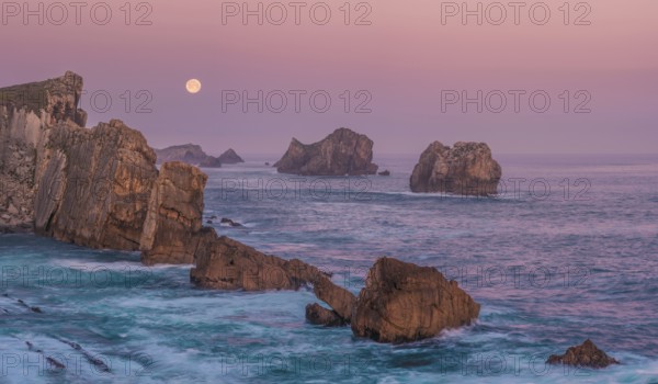 A serene view of rugged sea stacks and cliffs bathed in soft dawn light at Arnia Beach, Costa Quebrada, with the full moon glowing over the tranquil Atlantic horizon