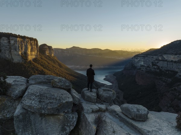 A lone figure stands on rocky cliffs, gazing over the Panta de Sau Reservoir in Catalonia, Spain, as the sun sets, painting the landscape in warm hues and shadows