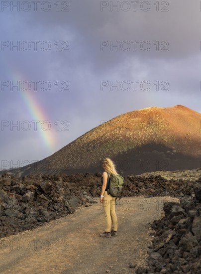 A woman with a backpack stands on a rocky path in Timanfaya National Park, Lanzarote. A vibrant rainbow arcs over the volcanic landscape under a dramatic sky