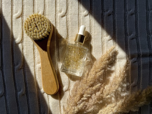A wooden brush and serum cosmetic bottle with golden serum rest on textured fabric, enhanced by soft lighting in the bathroom. Dried pampas grass adds an organic touch to the skincare composition