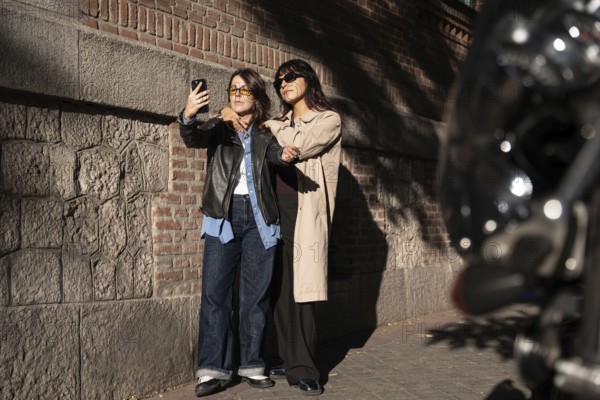 A multiethnic lesbian couple poses for a joyful outdoor selfie, exuding pride and love. The duo stands close, capturing a moment of happiness and unity