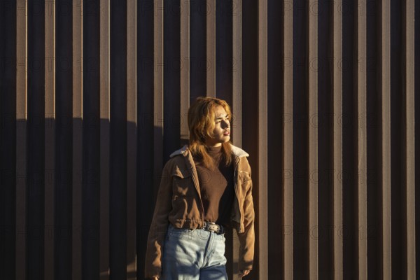 A Mixed-race woman stands against a vertical wooden wall, basking in warm sunlight She wears a casual brown jacket and jeans, embodying a relaxed, pensive mood