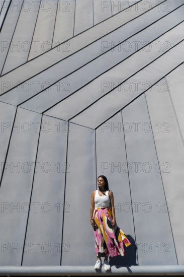 A woman in a vibrant, colorful outfit stands against a backdrop of sleek, modern architecture. Her attire creates a striking contrast with the geometric urban background