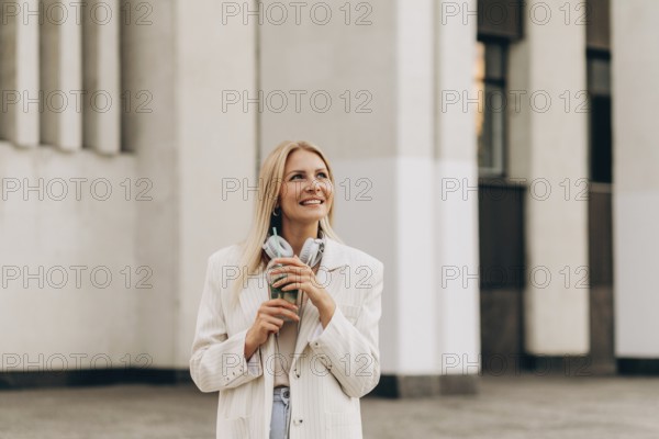 A smiling woman stands outside in an urban setting, holding a drink and headphones. She wears a light blazer, embracing the ambiance of the cityscape