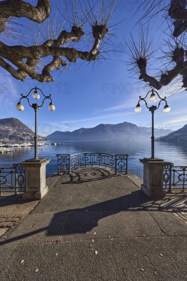 Lake, lakeside promenade, lanterns, trees, railings, mountains, haze, shadow, backlight, Lake Lugano, Lugano, Lugano district, Canton Ticino, Switzerland
