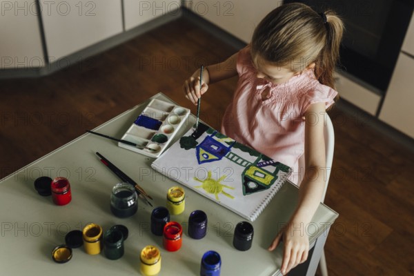 A child, dressed in a pink top, is intently painting a vibrant scene of houses and a sun using watercolors at a table. Several paint jars and brushes are scattered around