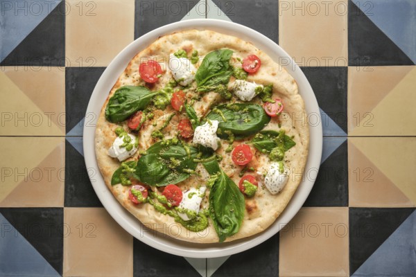 Top view of a delicious pizza with pesto, cherry tomatoes, basil and stracciatella cheese, served on a vibrant, geometrically tiled table