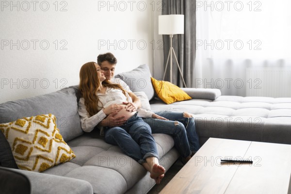 A young couple sits lovingly embraced on a gray sofa, hands gently resting on the woman's pregnant belly, reflecting a heartfelt anticipation of welcoming their baby into the world