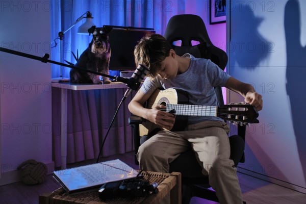 A young boy plays an acoustic guitar in a modern room with colorful lighting. A dog sits nearby on a desk, creating a cozy and creative atmosphere at home