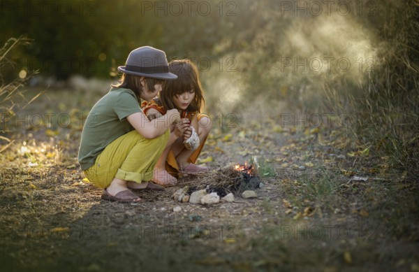 Two children sitting closely outdoors by a small campfire, surrounded by nature. They are focused on the fire, showcasing a sense of adventure and childhood exploration