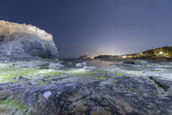 Tranquil Mediterranean shoreline on the Costa Blanca with rocky cliffs and calm sea under a starry night sky. Lights from distant shores reflect on the serene water surface