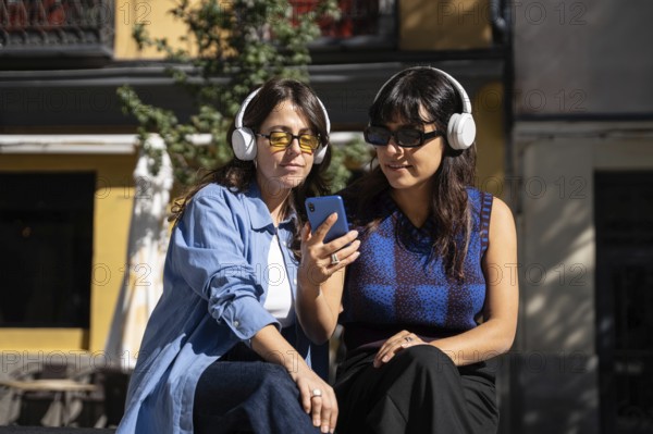A multiethnic lesbian couple sits outdoors, listening to music on headphones and sharing a smartphone. They embrace diversity and pride in their relationship