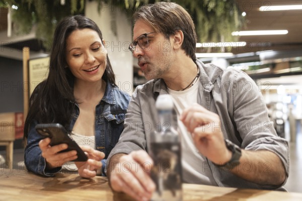 A couple sits at a wooden table, engaged in a lively conversation while sharing something on a smartphone. They appear relaxed and happy in a warmly lit indoor setting