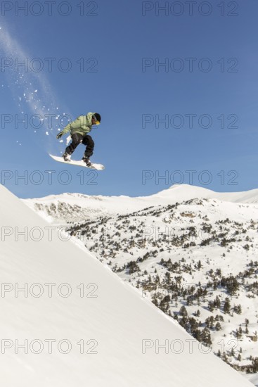 A snowboarder performs an airborne trick over a snow-covered mountain, capturing the essence of freeride and backcountry snowboarding. The fresh powder enhances the thrill