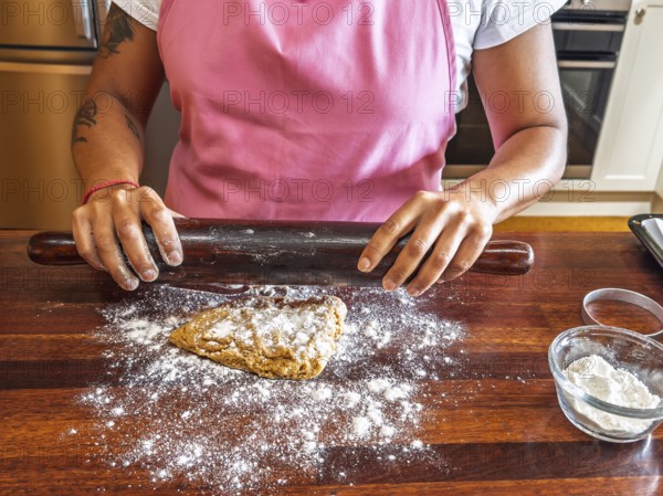 A woman in a pink apron uses a dark rolling pin to flatten dough on a floured wooden counter. A small glass bowl filled with flour is nearby, adding to the baking process