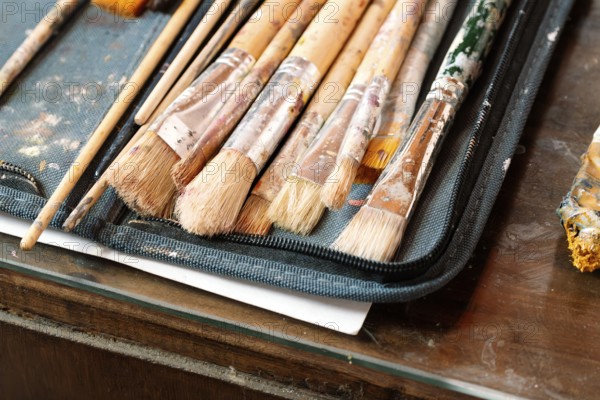 A close-up of well-used paintbrushes with dried paint resting in a fabric case on a wooden table. The worn textures and colorful bristles reflect the creative process of an artist