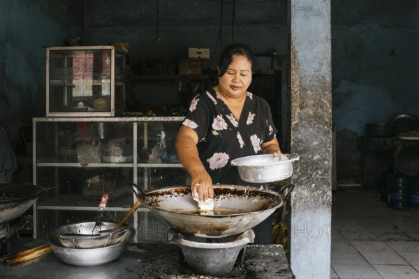 A woman prepares traditional food in a rustic kitchen of Bali, focusing intently as she cooks over a large wok. The indoor setting is simple, with an authentic local atmosphere