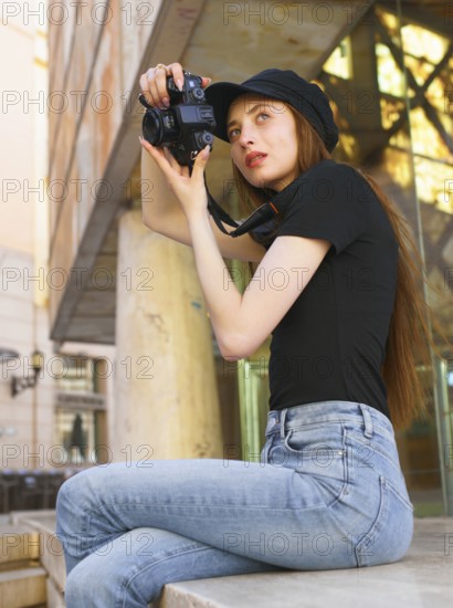 A woman in casual attire takes photos with a professional camera in a city setting. Her focused expression and stylish look reflect her passion for photography and urban exploration