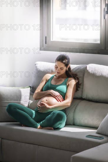 A pregnant woman peacefully sitting on a sofa in a sunlit room, dressed in a comfortable green yoga outfit, embracing the tranquility of pregnancy wellness and relaxation