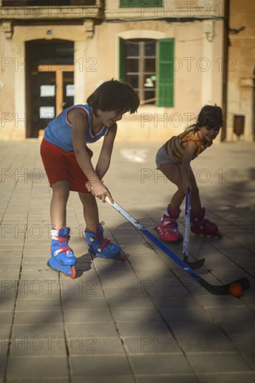 Two children play street hockey on a sunny day, wearing roller skates in an urban setting. The scene captures a fun and active pastime for kids