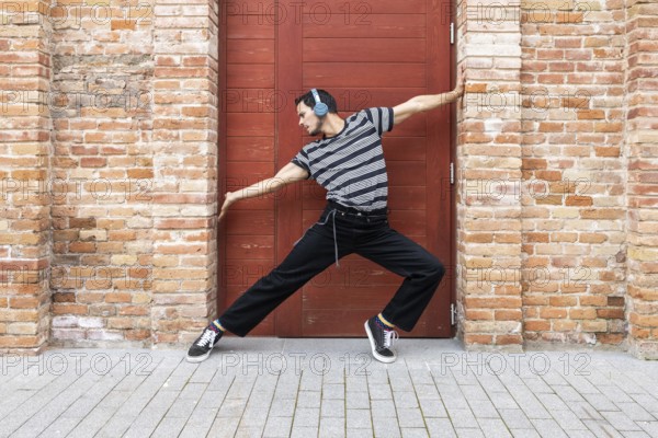 A cuban dancer exudes creativity and style, striking a dynamic pose against a vibrant red door framed by rustic brick. His casual attire and headphones reflect a modern vibe