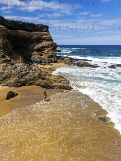 From above drone view of a lone woman walks on the sandy shore of Tindaya Beach in Fuerteventura, amidst rugged cliffs and foamy sea waves
