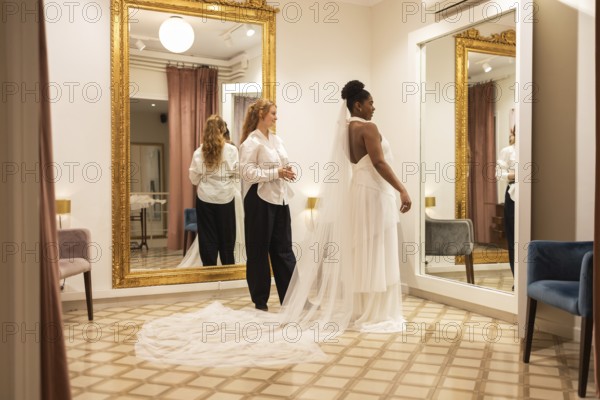 A bride admires her reflection in an elegant boutique fitting room. An assistant helps with her gown as they stand in front of large gold framed mirrors