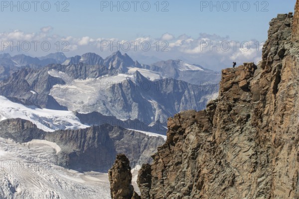 A hiker stands atop a rugged cliff in Zermatt, Switzerland, admiring the panoramic summer view of the majestic Alps, with glaciers and rugged mountain ranges extending into the horizon