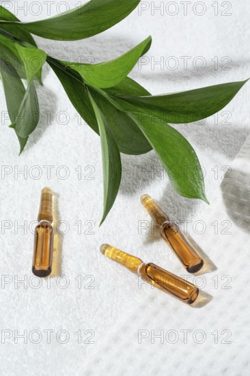 Close-up view of cosmetic ampoules with anti-aging serum, stylishly presented on a textured white towel and accented with vibrant green leaves for enhanced natural appeal