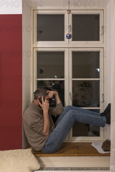 Young man sitting on the windowsill while talking on the phone, Mecklenburg-Vorpommern, Germany