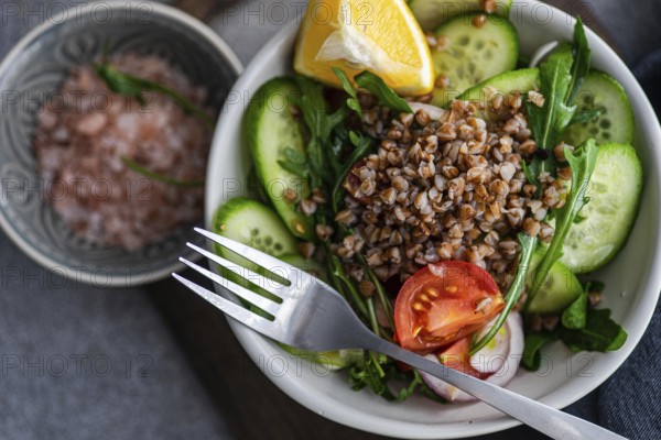 A nutritious lunch is prepared with fresh rocket, chopped oregano, cherry tomatoes, cucumber, Himalayan salt, red onion, steamed buckwheat, lemon, and organic olive oil