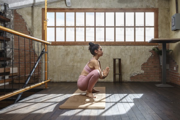 A woman wearing pink active wear is practicing yoga indoors on a sunny day. She is in a squatting pose, surrounded by rustic, wooden interior design elements