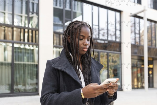 A young African American woman with braids, dressed in a business coat, is using a smartphone while standing outdoors near a modern building