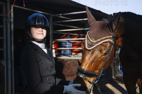 A teenage girl in classical dressage attire gently interacts with her bay horse. She wears a tailored black jacket and helmet, and the horse dons an ornate ear bonnet