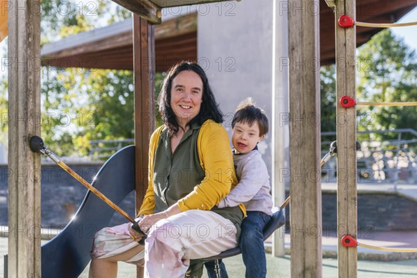 A joyful mother and child with Down syndrome are sitting together on a playground swing on a sunny day. The image captures a warm and happy moment with smiles and play on the equipment