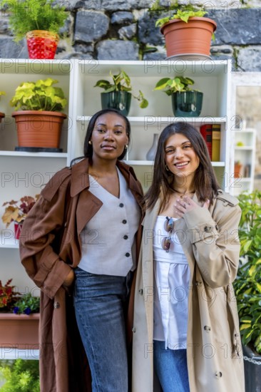 Two young businesswomen wearing stylish trench coats are posing confidently next to a shelf adorned with vibrant plants and decorative pots