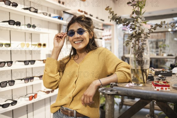 A cheerful Latin woman wearing stylish sunglasses poses in a trendy eyewear shop, showcasing a variety of fashionable frames. Her vibrant smile exudes confidence and style