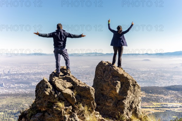 Two friends stand triumphantly on a mountain peak, facing a clear blue sky and expansive landscape, symbolizing freedom and adventure on a bright, sunny day
