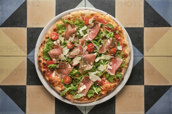 Overhead view of a whole pizza topped with prosciutto, arugula, and tomatoes, served on a vibrant, geometrically tiled surface
