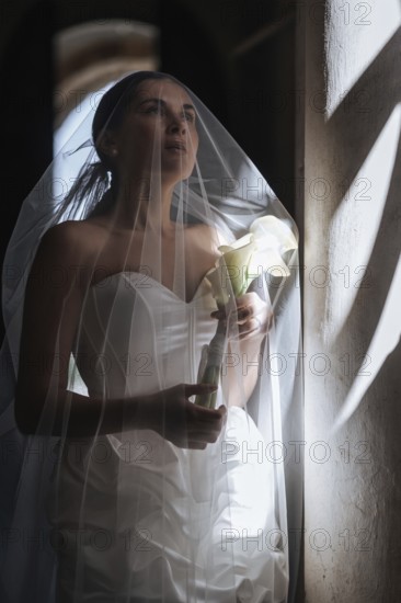 A bride in a stunning white gown stands gracefully inside an ancient villa in Treia, Macerata. The soft glow enhances the elegance of her veil and bouquet